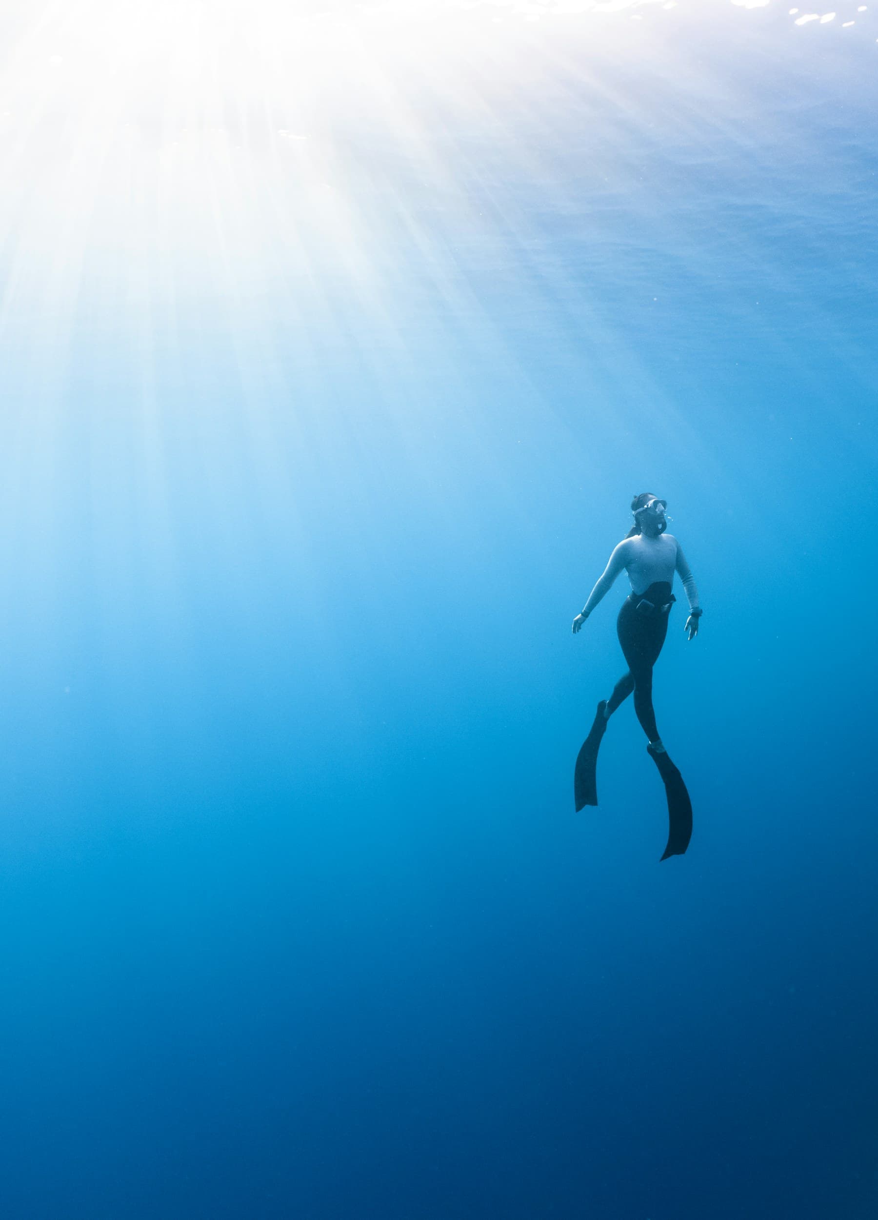 A freediver ascending toward sunlight at the surface of deep blue water.